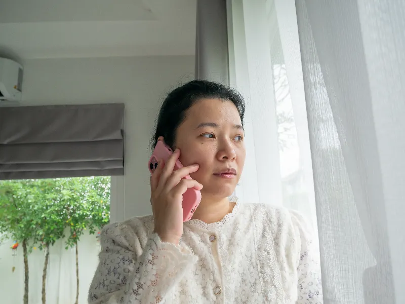 Woman in a white lace top talking on a pink phone while looking out a window with sheer curtains.