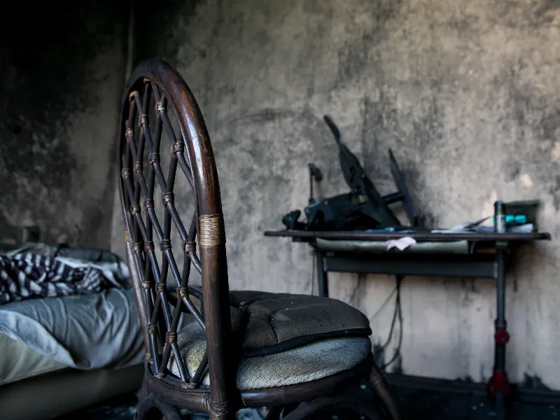 Worn wooden chair with woven backrest in front of a cluttered desk and bed in a dim room.