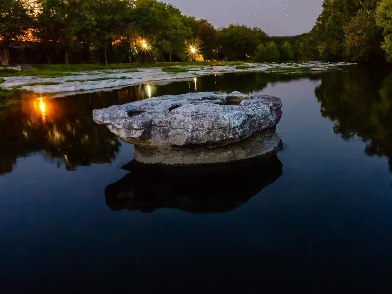 Large flat rock in calm river water at dusk with trees and lights in the background.