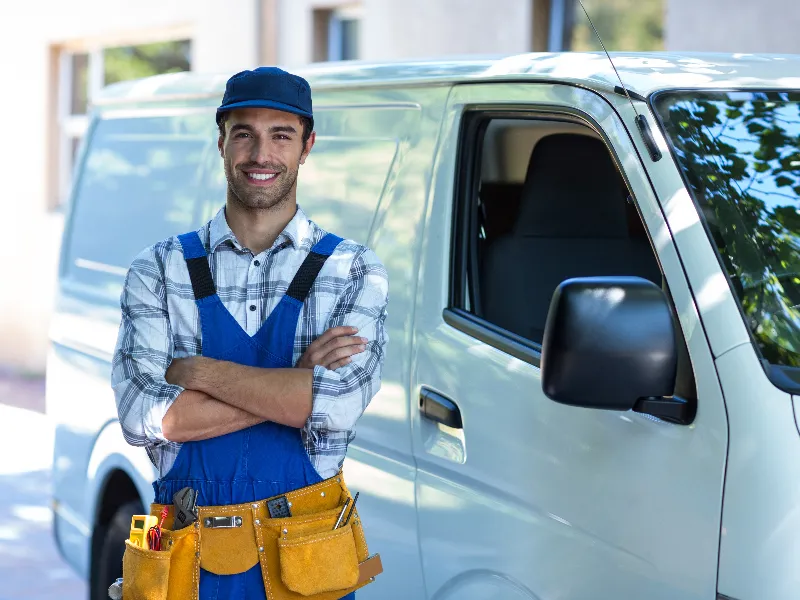 Smiling handyman in blue overalls and cap stands with arms crossed beside a white van.