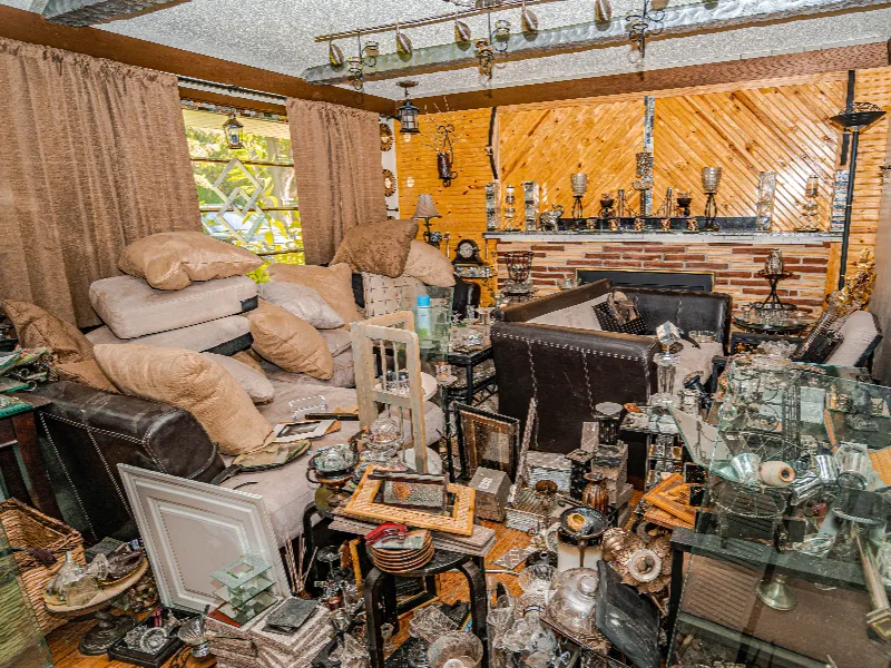 Cluttered living room filled with stacked cushions, glassware, picture frames, and various decorative items.