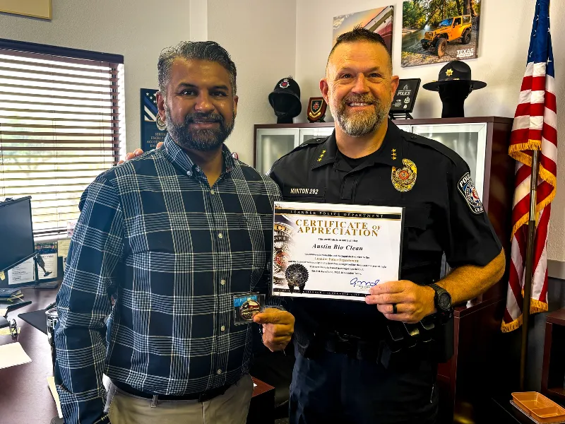 A man in a plaid shirt and a police officer hold a certificate and badge in an office with an American flag.